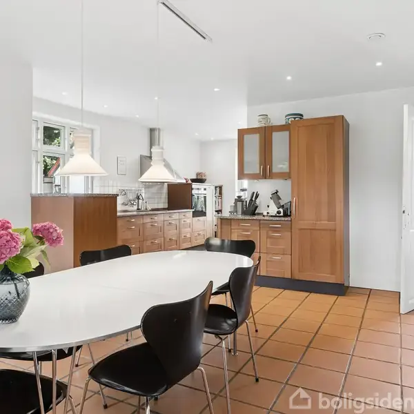 En moderne køkken-spisestue med ovalt hvidt bord med sorte stole. En vase med blomster står på bordet. Træskabe, komfur og to loftslamper udgør køkkenområdet. Pejs i højre hjørne.