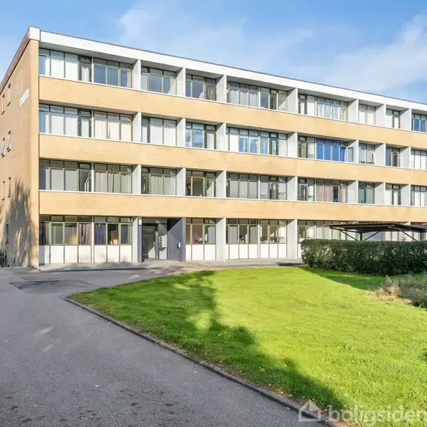 En treetagers, moderne boligblok med mange vinduer står stille. Den er omkranset af en asfalteret vej og grønne områder under en blå himmel.