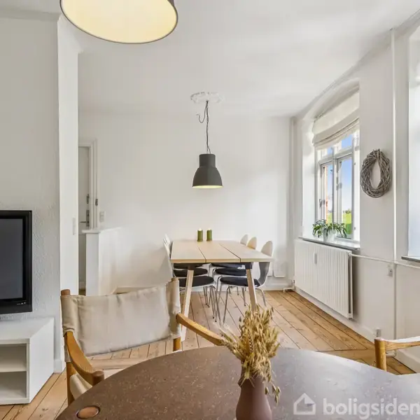 I en lys stue står en fladskærms-tv på et hvidt møbel. Et rundt bord med stole ses foran. Vinduet bagved lader sollys strømme ind, afslørende træernes grønne løv udenfor.