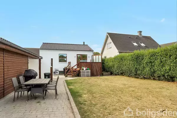 Et hvidt hus med en terrasse og trappe i en baghave. En havegrill og et bord med stole står på flisebelægning. Grønne hække omkranser græsplænen i et villakvarter.