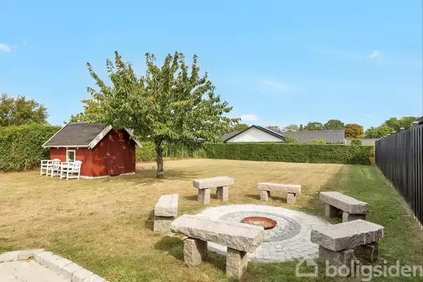 Et rødt haveskur står stille ved et træ, omgivet af en græsplæne og hække. En cirkel af stensæder omringer et bålsted, under en blå himmel.