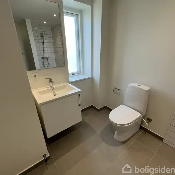Toilet står ved siden af et brusebad og en vask med et spejl i et moderne, lyst badeværelse med flisegulv og en lille vinduesåbning.