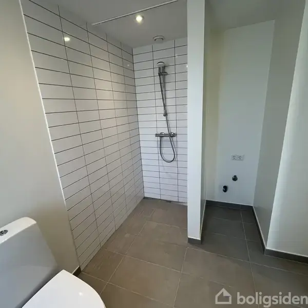Bruseniche med hvidt flisebelagt væg står i et moderne badeværelse. Der er en toilet i forgrunden og lyse vægge omkring, hvilket skaber et minimalistisk udseende.