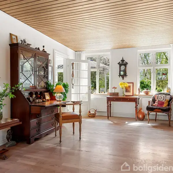En lys stue med trægulv og loft, indeholder antikke møbler, planter og dekorative elementer. Store vinduer og en glasdør tillader naturligt lys. Vægur hænger på væggen.