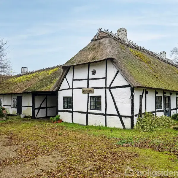 Bindingsværkshus med stråtag står i natur omgivet af træer og buske. Et skilt på facaden siger "Frendeløst", og huset har sort-hvide detaljer og flere små vinduer.