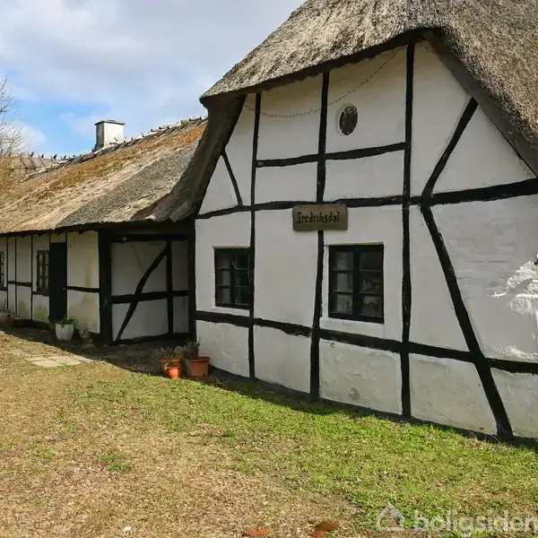 Bindingsværkshus med hvidkalkede vægge og sort træværk. Skiltet siger "Solhøjsbak". Huset har stråtag og flere små vinduer. Omgivet af ubeskrevet natur med en klar himmel.