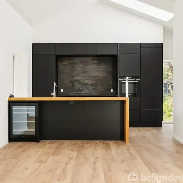 Et moderne køkken med sort kabinet, hvor indbyggede apparater og en køkkenø står i fokus. Køkkenøen har en håndvask og en integreret vinkøler. Rummet er lyst med trægulve.