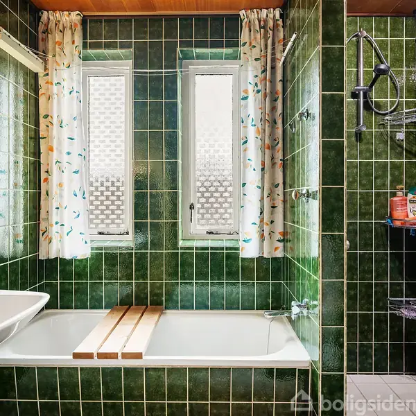 Badeværelse med grønne fliser, badekar og bruseområde. Badekarret har en trærist, og der hænger gardiner ved de to vinduer. Shampoo og sæbe opbevares på hylder i bruseområdet.