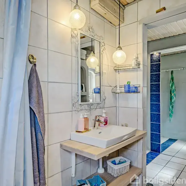 Håndvask med rektangulært spejl hænger på en hvidklinket væg. To loftlamper lyser området op. Hylder med toiletartikler ved siden af. Blå fliser danner en arkitektonisk ramme ind til bruseren.