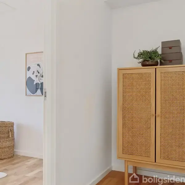 Et vægskab af flet står i en lys entré, dekoreret med planter og æsker på toppen. Til venstre ses et soveværelse med en flettet kurv ved siden af sengen.