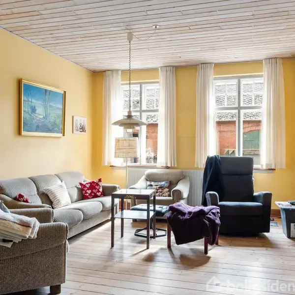 Stue med to sofaer, et sofabord og en lænestol i et rum med gule vægge og trægulv. Vinduerne har lyse gardiner, en lampe hænger fra loftet. Ingen tekst.