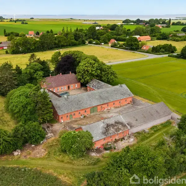 En gammel, murstenslade står ubenyttet omgivet af frodige marker og spredte træer i et landligt landskab. Adskillige mindre bygninger befinder sig i nærheden langs en landevej.