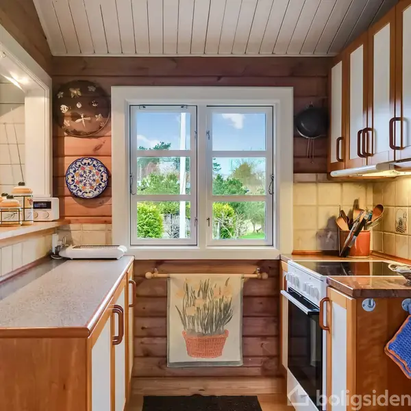Et køkken med træpaneler og hvid loft, indeholder en komfur med ovn til højre samt farverige håndklæder. Et vindue med udsigt til en grøn have centralt placeret. En hylde med blomster til venstre.
