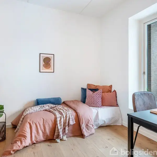 En enkeltseng med farverige puder og tæppe står i et lyst værelse med trægulv. Ved siden af er en reol med planter, og et skrivebord ved vinduet.