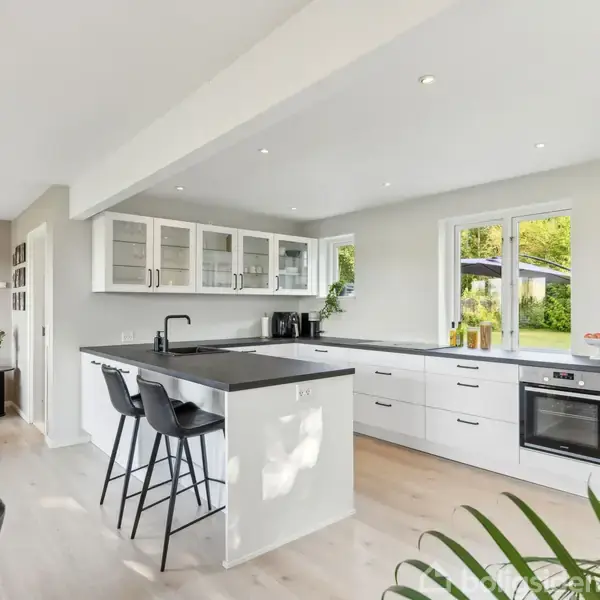 Et moderne køkken med hvide skabe og sorte bordplader. To barstole står ved en køkkenø. Rummet er lyst med store vinduer ud til en grøn have. En stue ses i baggrunden.