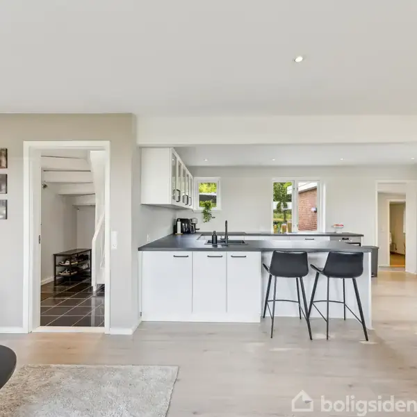 Et hvidt køkken med sort bordplade, to sorte barstole ved en ø. Til venstre ses en døråbning mod en gang og trappe, omgivet af lyse vægge og træmøbler.
