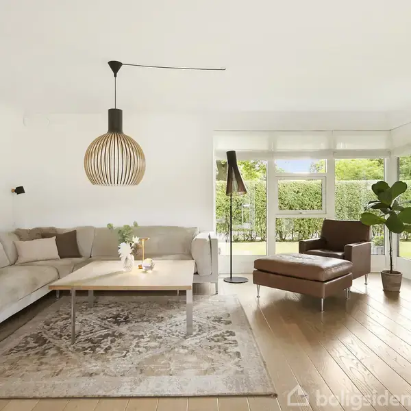En moderne stue med en lys sofa og sofabord på et tæppe. En stor lampe hænger over bordet. Der er en lænestol ved vinduer, der ser ud til haven.