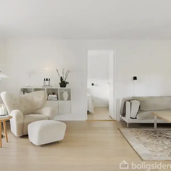 En stue med en lænestol og sofabord står på et lyst trægulv. Rummet har lyse vægge og indeholder en sofa, lampe og plantereol. En dør fører til et andet rum.