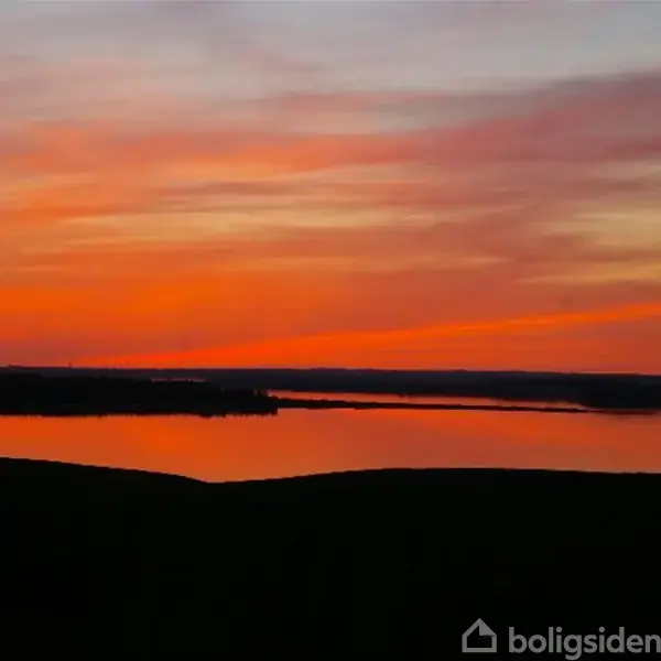 Sø reflekterer en intens orange og rød solnedgang, mens mørke silhuetter af land omkranser horisonten. Himlen er farvestrålende med bløde skyformationer.