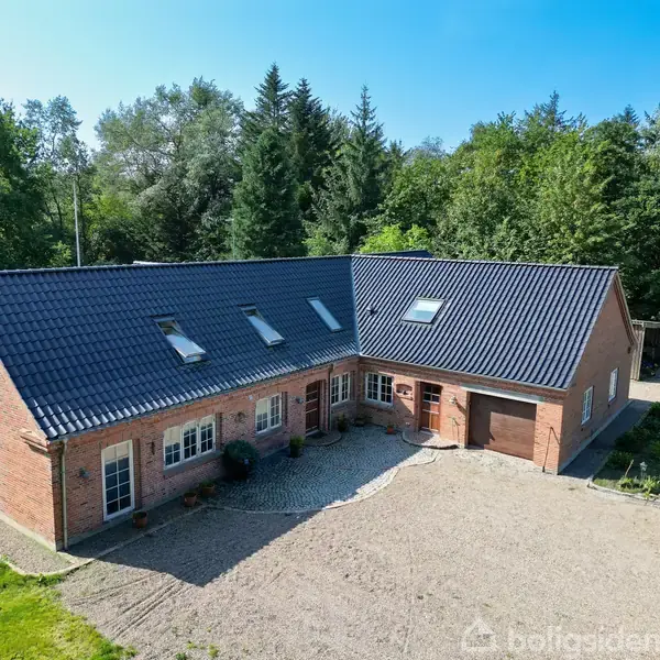 Et murstenshus med sort tegltag står på en grusbelagt plads omgivet af skovklædte træer. Husets indgangsparti har flere vinduer og en garage i højre side.