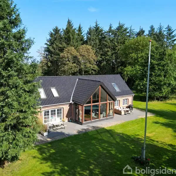 En rummelig villa med store vinduespartier står på en grøn græsplæne omgivet af høje træer. Huset har en mørk tagkonstruktion og en stor terrasse med havemøbler.