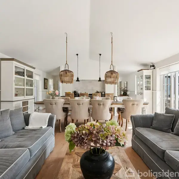 Grå sofaer står omkring et træbord med en vase fyldt med blomster. I baggrunden er der et moderne køkken med høje barstole og hængende lamper.