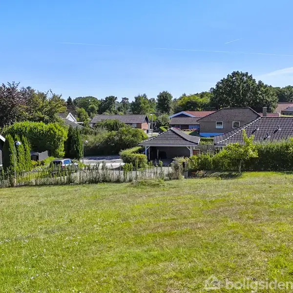 Græsplæne strækker sig over en bakke, omgivet af huse med mørke tagsten i en forstad. Træer og buske kantet de velholdte haver under en blå himmel.