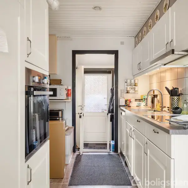 Køkken med hvide skabe og apparater står langs væggene. En dør i baggrunden fører ud til en anden del af boligen, mens en tæppebelagt gangvej ligger centralt.