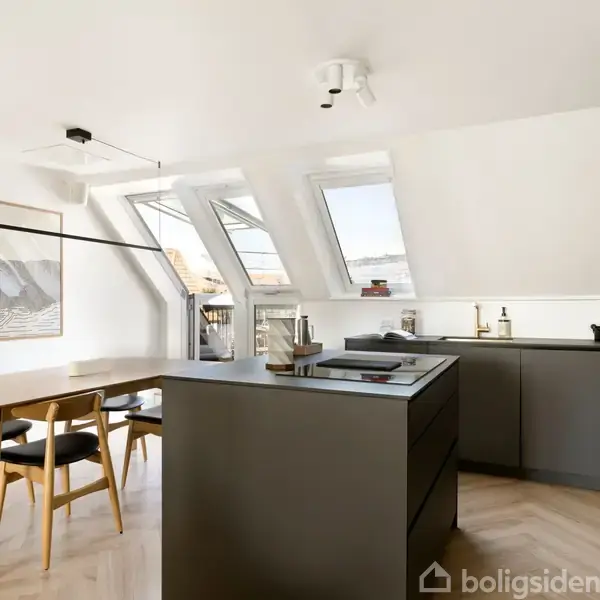 Køkken i moderne design med sort køkkenø, plader på væggene, spisebord med stole. Store skråvinduer giver lys, og hvide vægge skaber en lys atmosfære. Ingen tekst til stede.