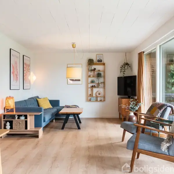 Stue med en blå sofa ved siden af et lille bord. To stole står overfor, med store vinduer til højre. Plakater hænger på væggen, og en lampe oplyser rummet.