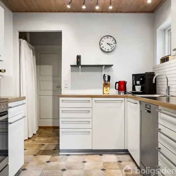 Et moderne køkken med hvide skabe. En ovn står til venstre. På bordet er der knive, elkedel og kaffemaskine. En hvid væg med ur og et lille vindue ses i baggrunden.