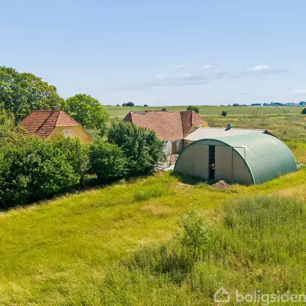 En grøn buet lade står stille mellem to ældre bygninger med røde tegltage, omgivet af højt græs og træer, midt i et åbent, landligt landskab under en lys himmel.