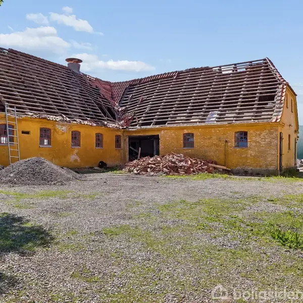 Gult murstenshus med delvist fjernet tag, omgivet af byggematerialer og en stige op ad facaden, i en gårdsplads med grus og græs, under en blå himmel med skyer.