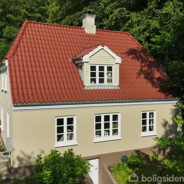 Et hvidt hus med rødt tegltag står stille i en skovklædt have. Huset har store vinduer og et centralt kvistvindue. Terrasse og buske omgiver boligen.