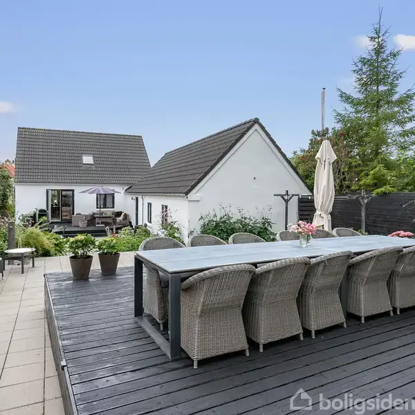 Spisebord med ni kurvestole står på en træterrasse. I baggrunden ses en hvid bygning med mørkt tag og terrasseområde. Omgivelserne indeholder planter og et hegn.