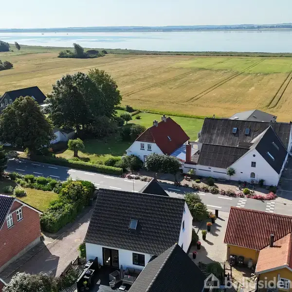 Flere huse står stille i en lille by med grønne haver. Omkring dem strækker marker sig mod en fjord i baggrunden. Sollys oplyser landskabet.