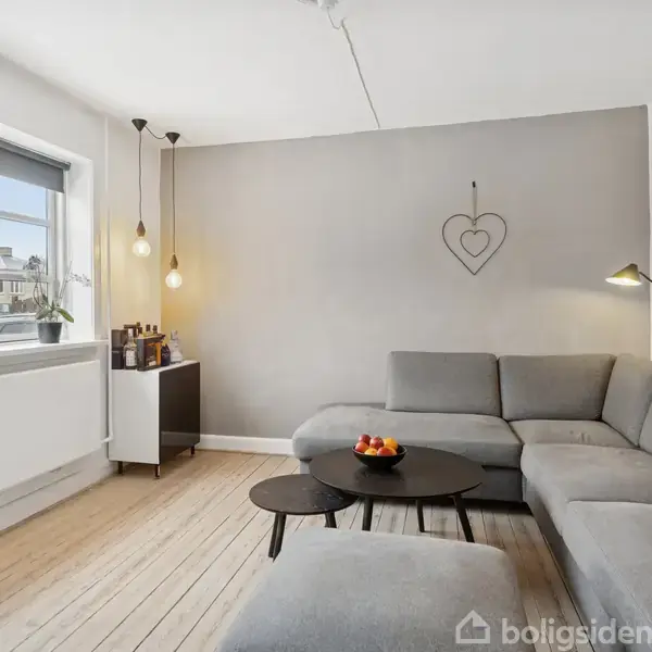 Gråt sofaarrangement står i stue med lyse trægulve, ved siden af et lille bord med en skål frugt. Vindue med planter slipper lys ind. To lamper hænger fra loftet.