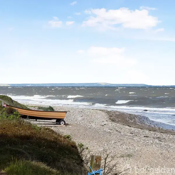 En jolle står på en trailer ved en stenstrand. Vinden skaber bølger på havet. Kystlinjen strækker sig i baggrunden under en blå himmel med spredte skyer.