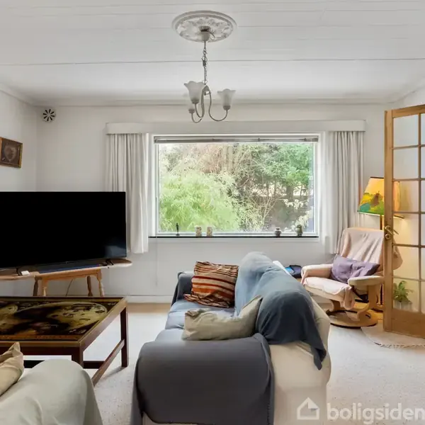 Stue med sofa og puder, et bord med et mønster, fladskærms-tv og et stort vindue med udsigt til en have. Der er også en lænestol, lampe, og franske døre.