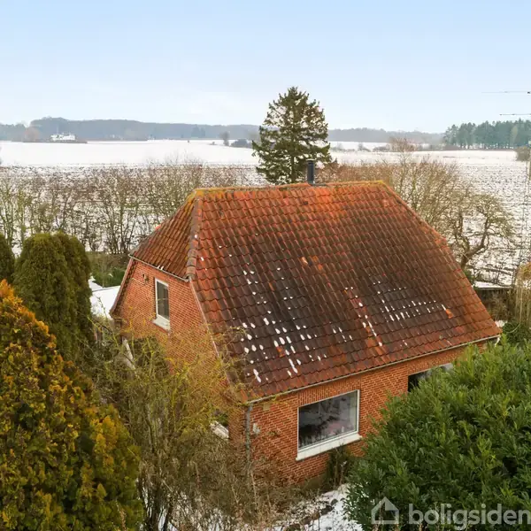 En rød murstenshus med et rødt tegltag står omgivet af sneklædte træer. Baggrunden viser åbne marker dækket af sne, under en klar blå himmel.