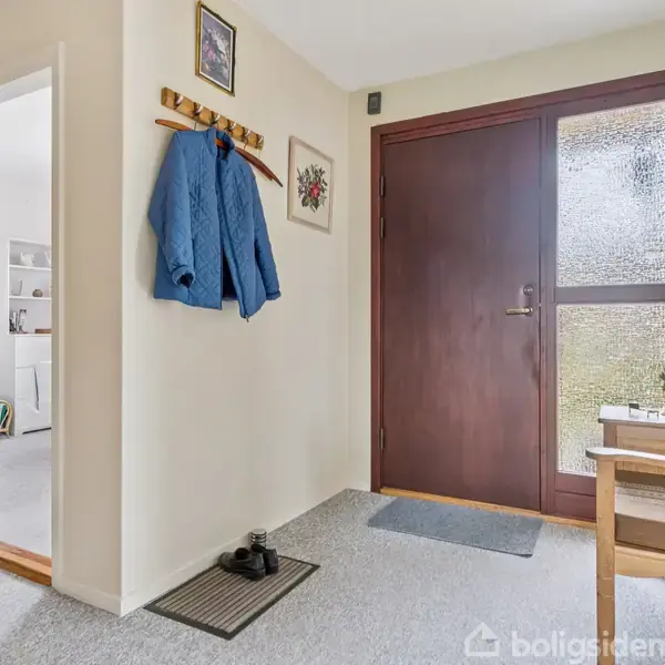 En gang med en trædør til højre, hvorpå der hænger en frakke på en knagerække. Ved siden af står en bænk med lysestager. Til venstre er en åbning til et andet rum.