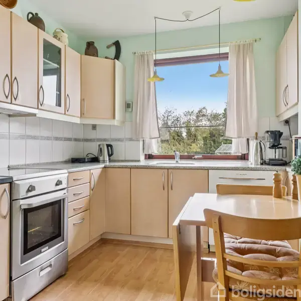 Køkkenbord med en træstol står foran et lys træskabinet. Et vindue med hvidt gardin viser udsigt til træer udenfor. Lyseblå vægge omkranser rummet. Ingen tekst.