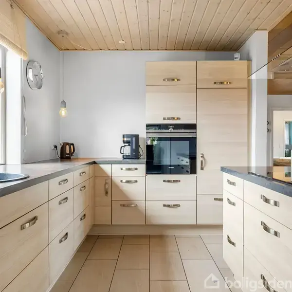 Et moderne køkken med lyse træskabe og grå bordplader. En kaffemaskine og elkedel står på bordet; en ovn er indbygget i skabene. Lyset strømmer ind fra et stort vindue.