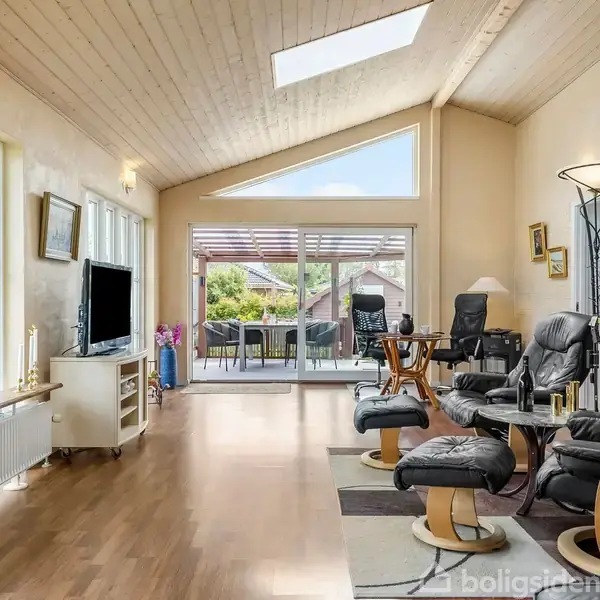 Stue med træloft og store vinduer; komfortable lænestole står nær et bord. Bag ved er en terrasse med udendørs møbler. Lyse omgivelser med moderne indretning.