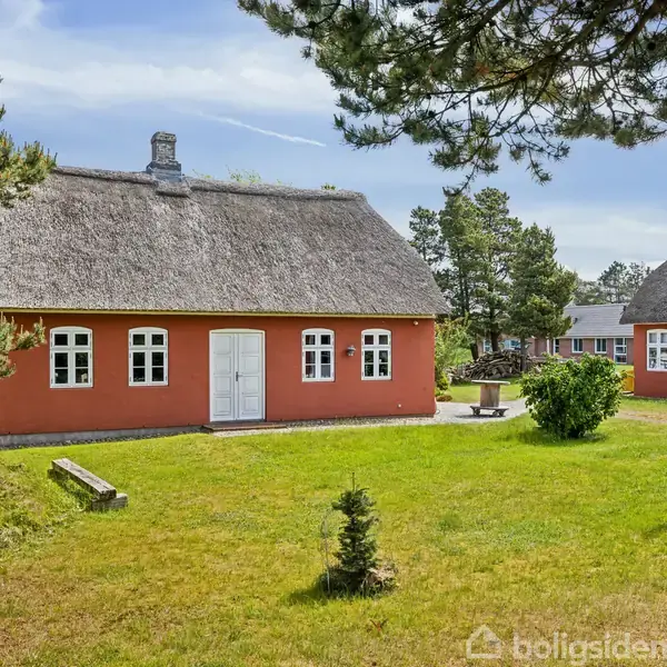 Et rødt bindingsværkshus med stråtag står stille på en velplejet græsplæne omgivet af høje træer. Et bordbænkesæt ses til højre, med et lignende hus i baggrunden.