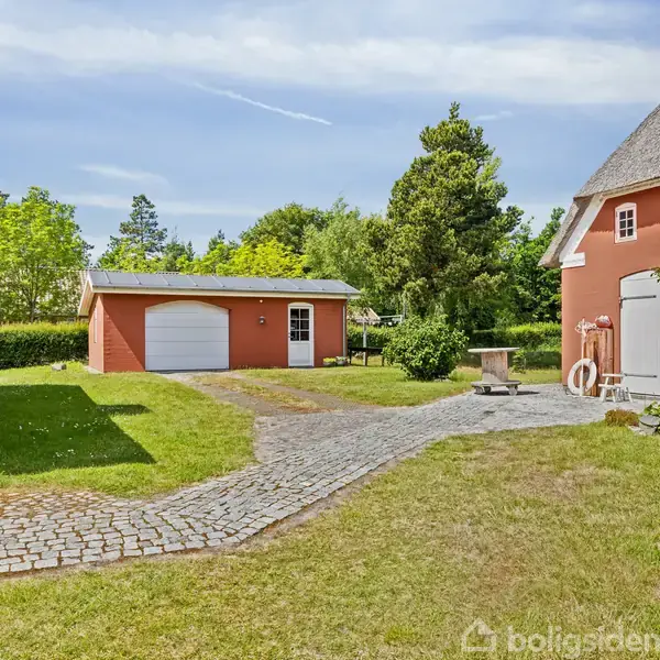 Røde bygninger med stråtag står stille; en midterste grusvej omkranset af grønne træer og græs; en garage ses i baggrunden med en hvid port.