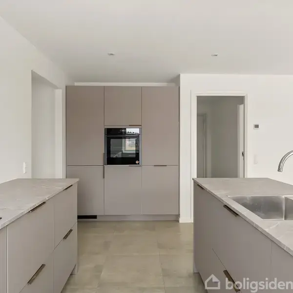 Køkken med moderne stil. Bordplader i grå, indbygget ovn og vask. Rummets vægge er hvide, og der er en åbning til en anden del af huset.