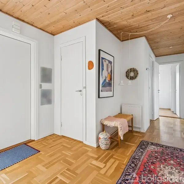 En entré med parketgulv og træloft har døre og skabe langs væggen. Et billede hænger på væggen over en bænk med en lys pude. En rød løber ligger på gulvet.