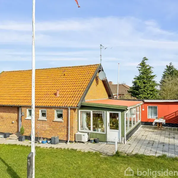 Rødt murstenshus med rødt tegltag står på en velholdt græsplæne. Et flisebelagt terrasseområde er foran huset, omgivet af grønne træer og buske i baggrunden. Flagstang til venstre.