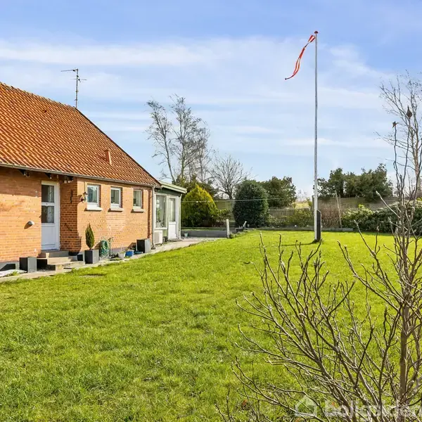 Et rødt murstenshus står stille ved siden af en velplejet græsplæne og en flagstang med flag i en have omkranset af buske og træer under en blå himmel.